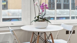 A room with a round table, two white chairs, a potted orchid, and a view of buildings through large windows. - Grove Heights Shoreditch  (London)