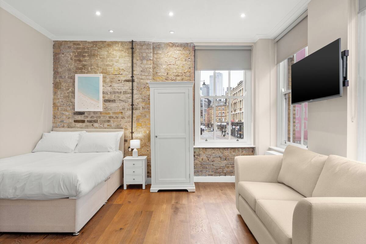 A bedroom with a bed, a nightstand, a dresser, a window with a city view, and a TV mounted on the wall.