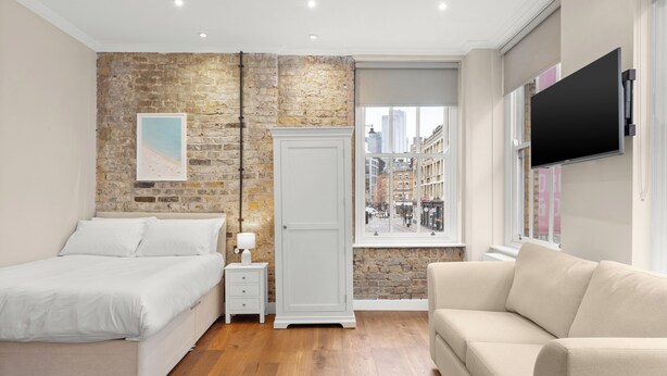 A bedroom with a bed, a nightstand, a dresser, a window with a city view, and a TV mounted on the wall. - Grove Heights Shoreditch  (London)