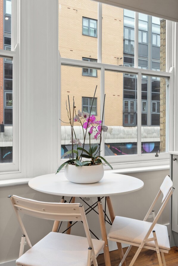 Una habitación con una mesa redonda, dos sillas, una orquídea en maceta y vistas a un edificio a través de amplias ventanas.