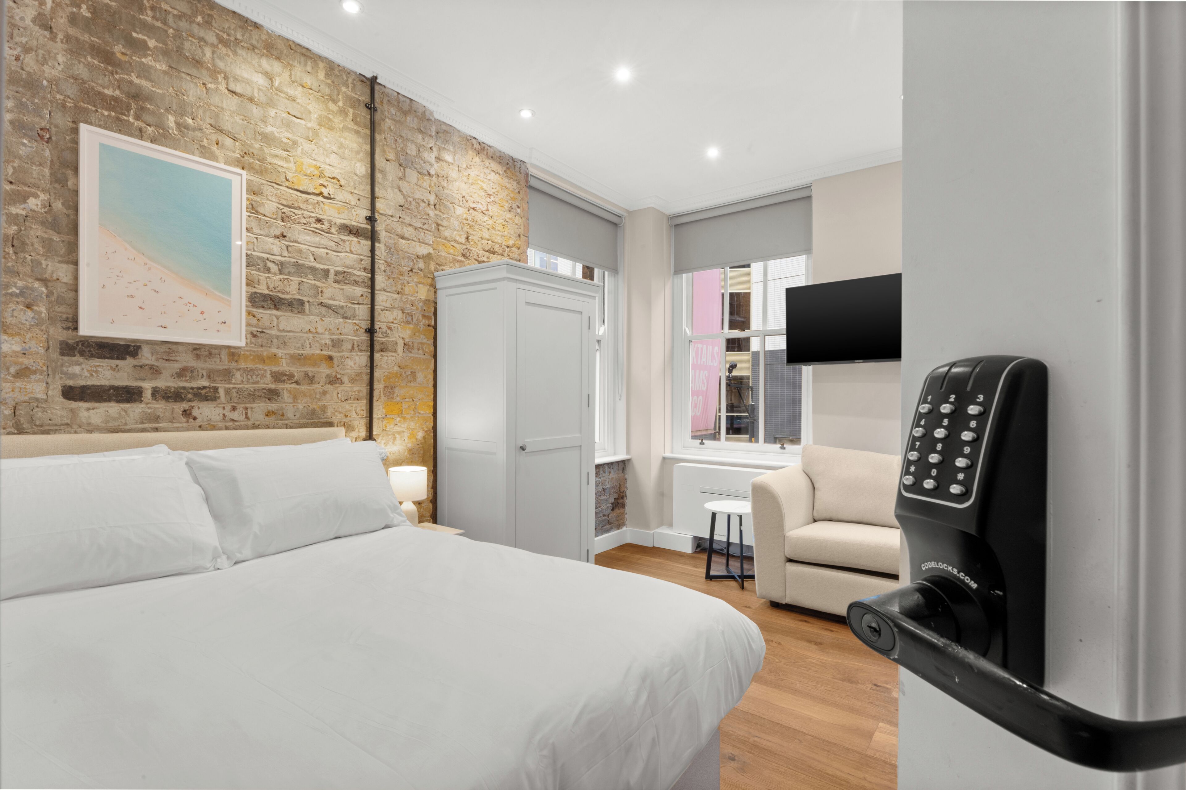 A hotel room with a brick wall, a bed, a wardrobe, a desk with a chair, and a TV mounted on the wall.