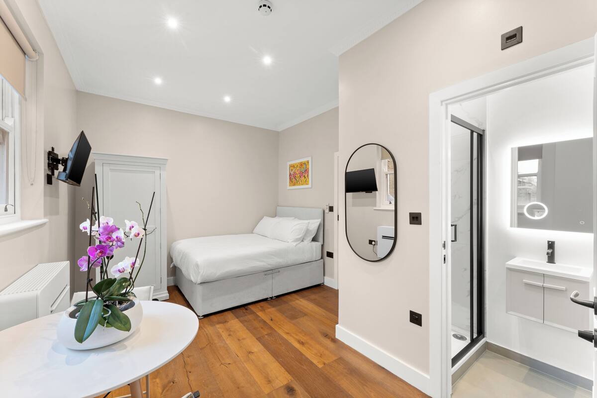 A modern bedroom with a bed, a round mirror, a white bedside table, and a potted orchid.