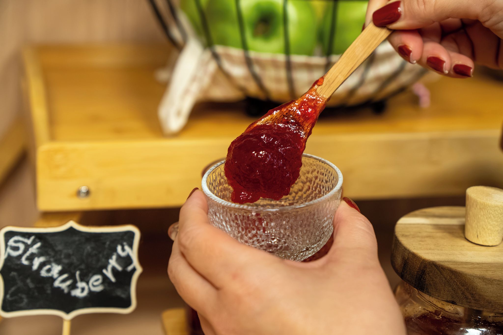 A person holding a spoon with a red substance, possibly jam, over a glass.