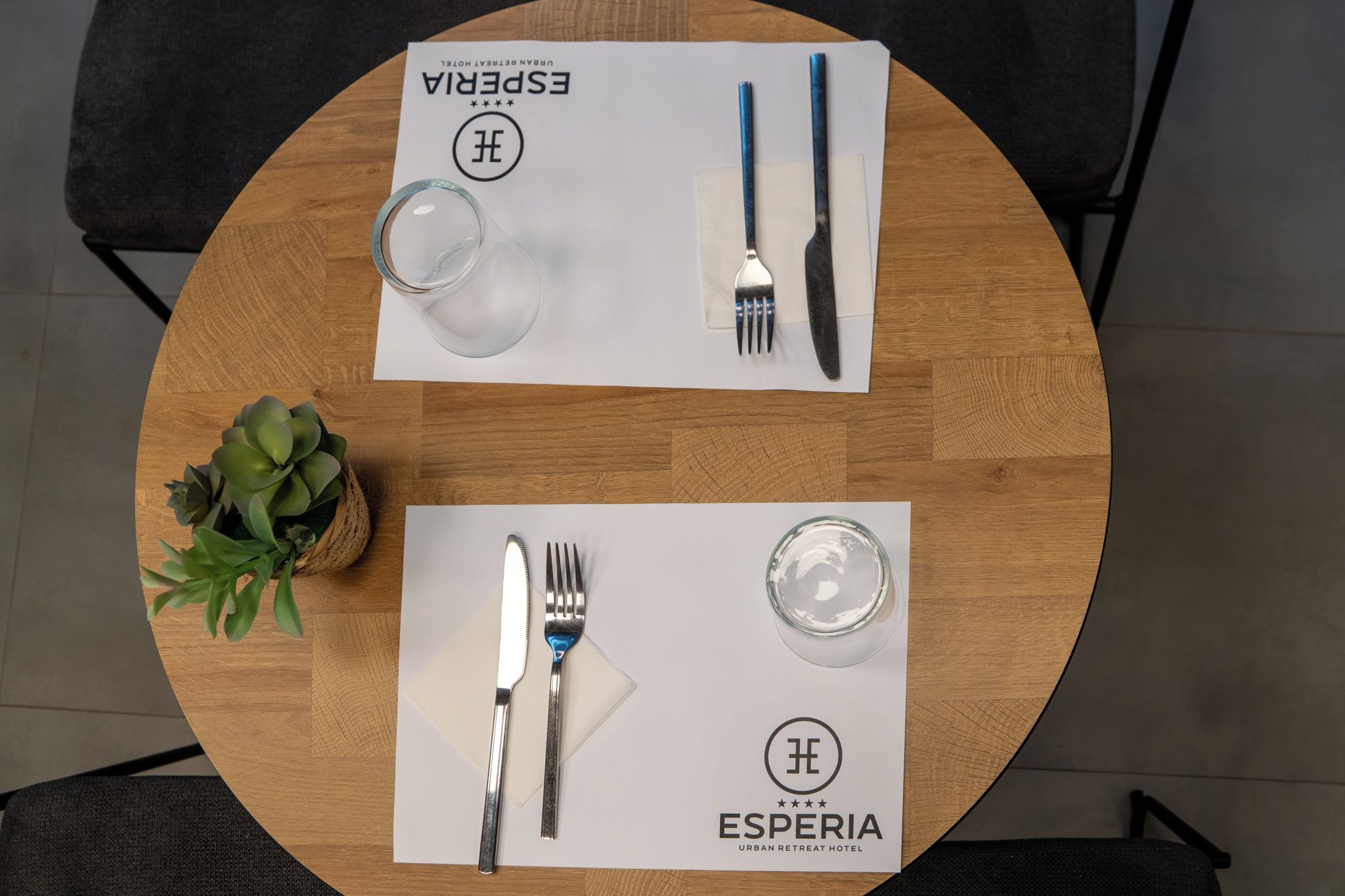 Una mesa de madera redonda con dos juegos de cubiertos y cristalería, cada uno con el logotipo 'ESPERIA', lo que indica una disposición para comer.