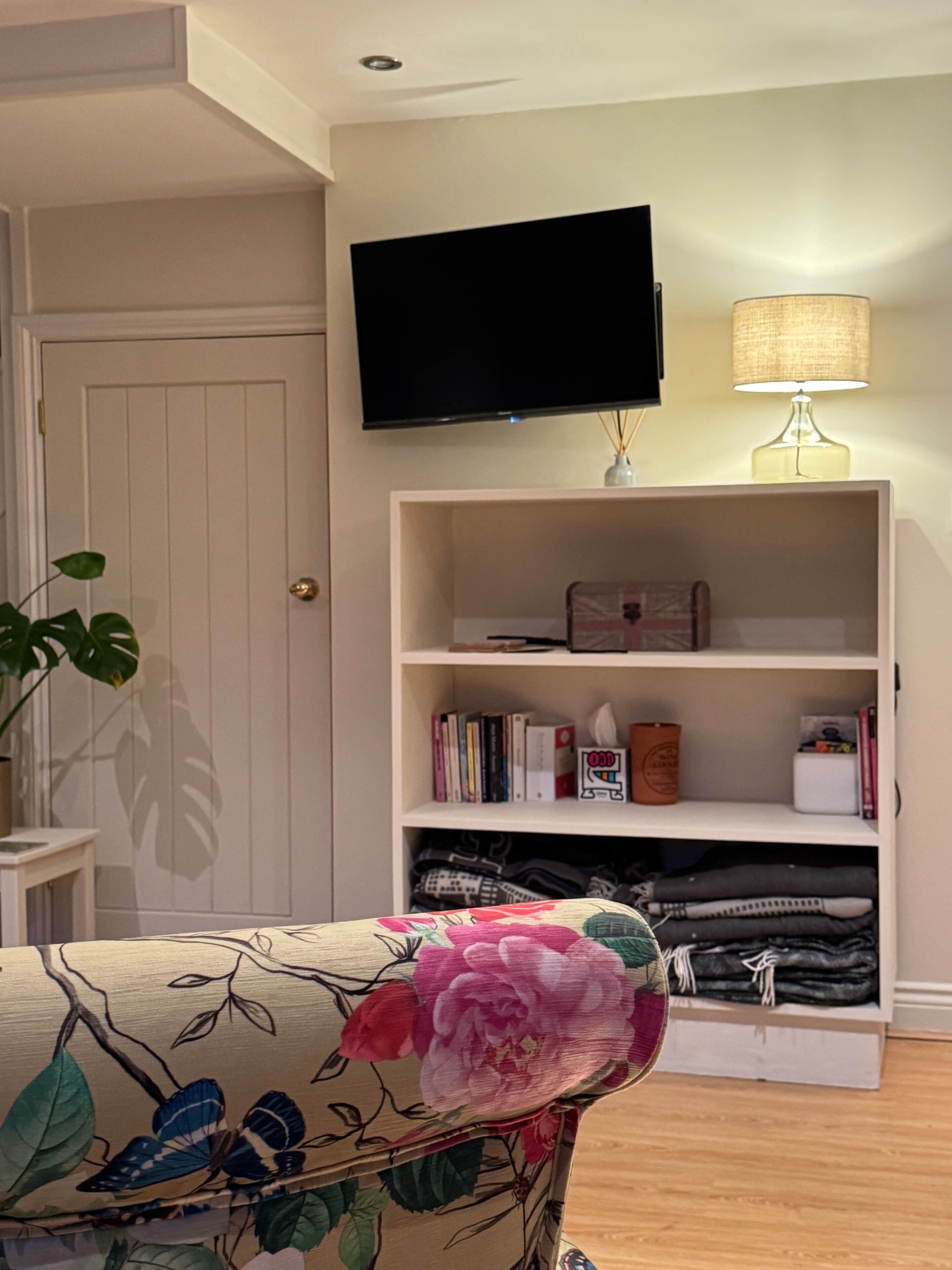 Ein Wohnzimmer mit an der Wand montiertem Fernseher, einer Bücherwand, einer Lampe und einem blumengemusterten Sessel.