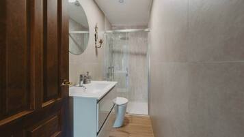 Un baño estrecho con puerta de madera, un lavamanos blanco, un inodoro y una ducha con cerramiento de vidrio.