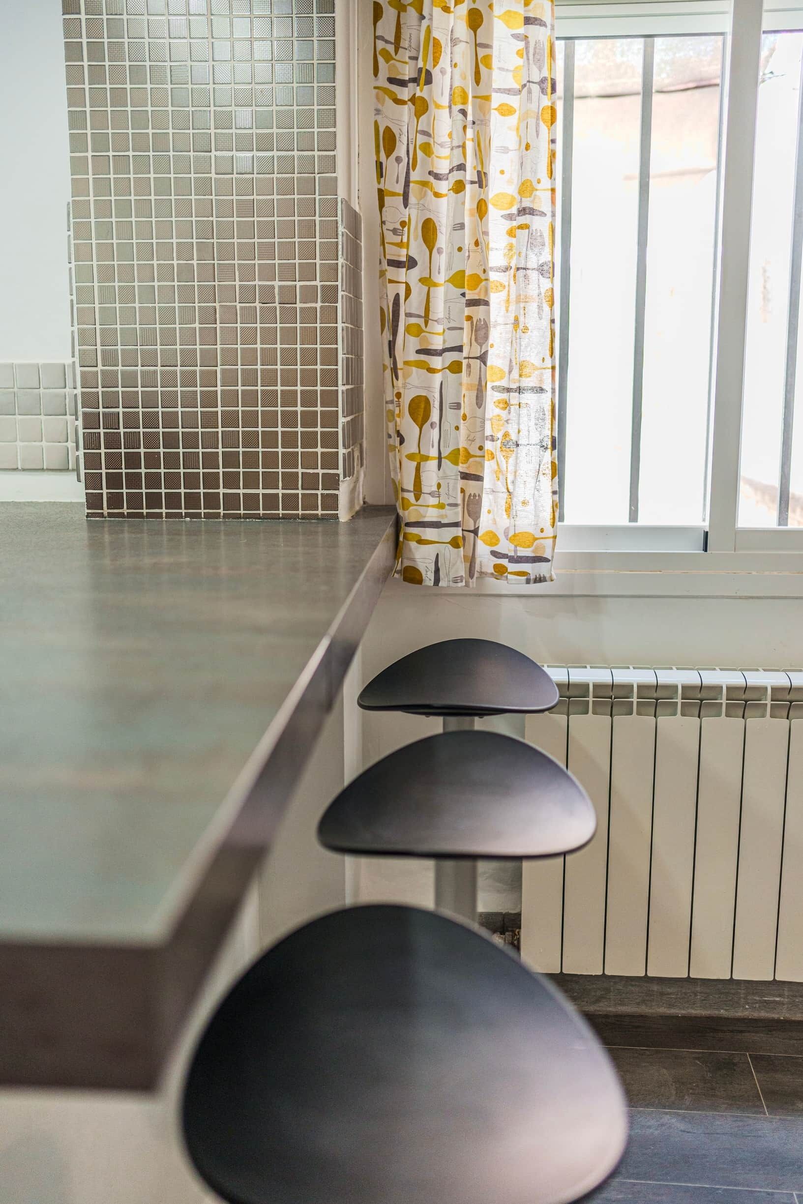 Una cocina con salpicadero de azulejos, un ventanal con cortinas y un taburete de bar.
