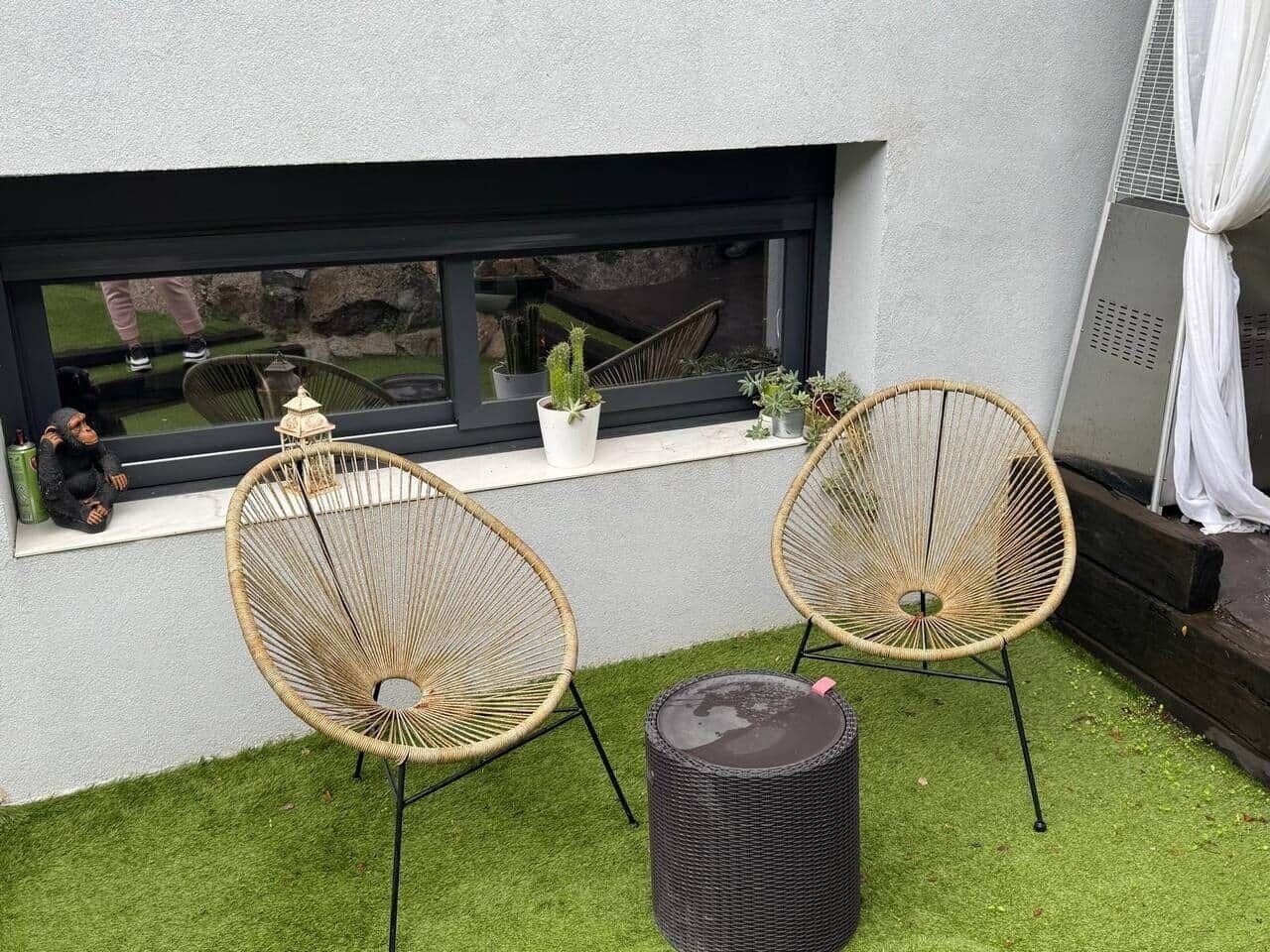 Dos sillas de mimbre, una mesita redonda y un cubo de basura negro sobre una zona cubierta de césped, con una ventana y plantas al fondo.