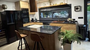 Ett modernt kök med en central köksö, barstolar, en inbyggd ugn och ett stort fönster med utsikt över en trädgård.