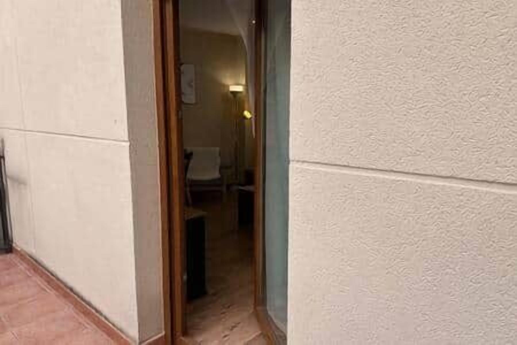 Un pasillo estrecho con una puerta de madera que da a una habitación. La habitación tiene suelo de baldosas y una silla.