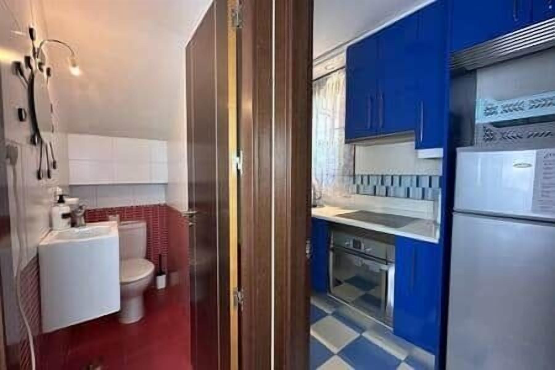 Een kleine badkamer met toilet, wastafel en douche. De aangrenzende ruimte is een keuken met blauwe keukenkastjes en een koelkast.