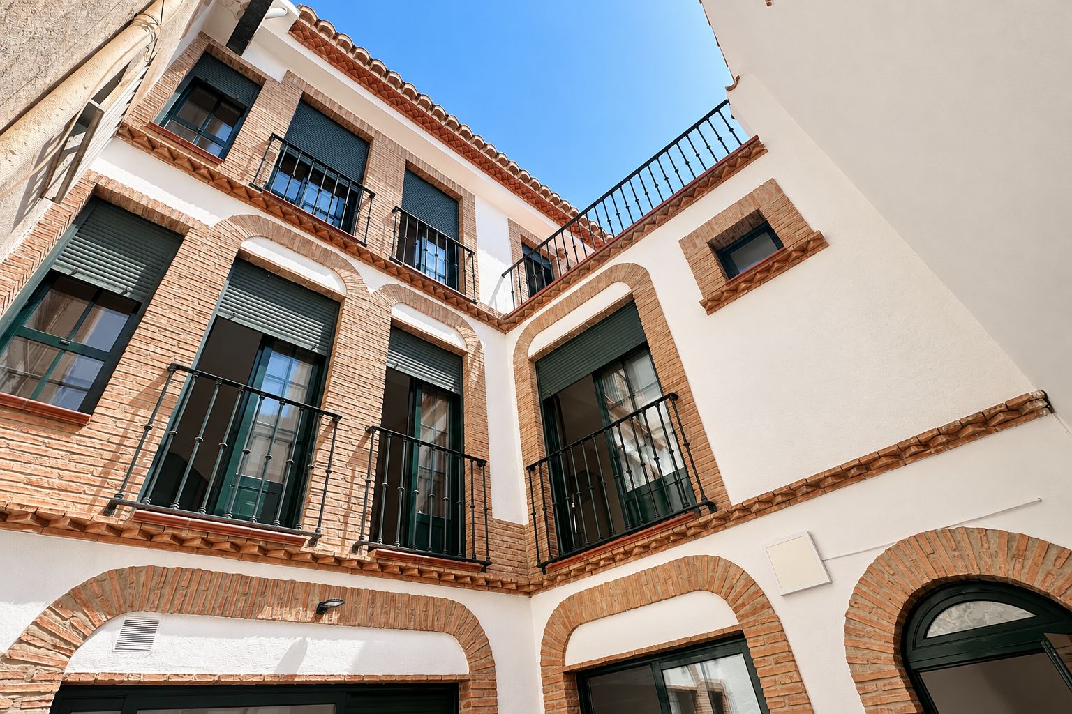 Un edificio de varios pisos con patio central, que presenta balcones con barandillas negras, ventanas de arco y una combinación de fachadas de ladrillo y enlucido.