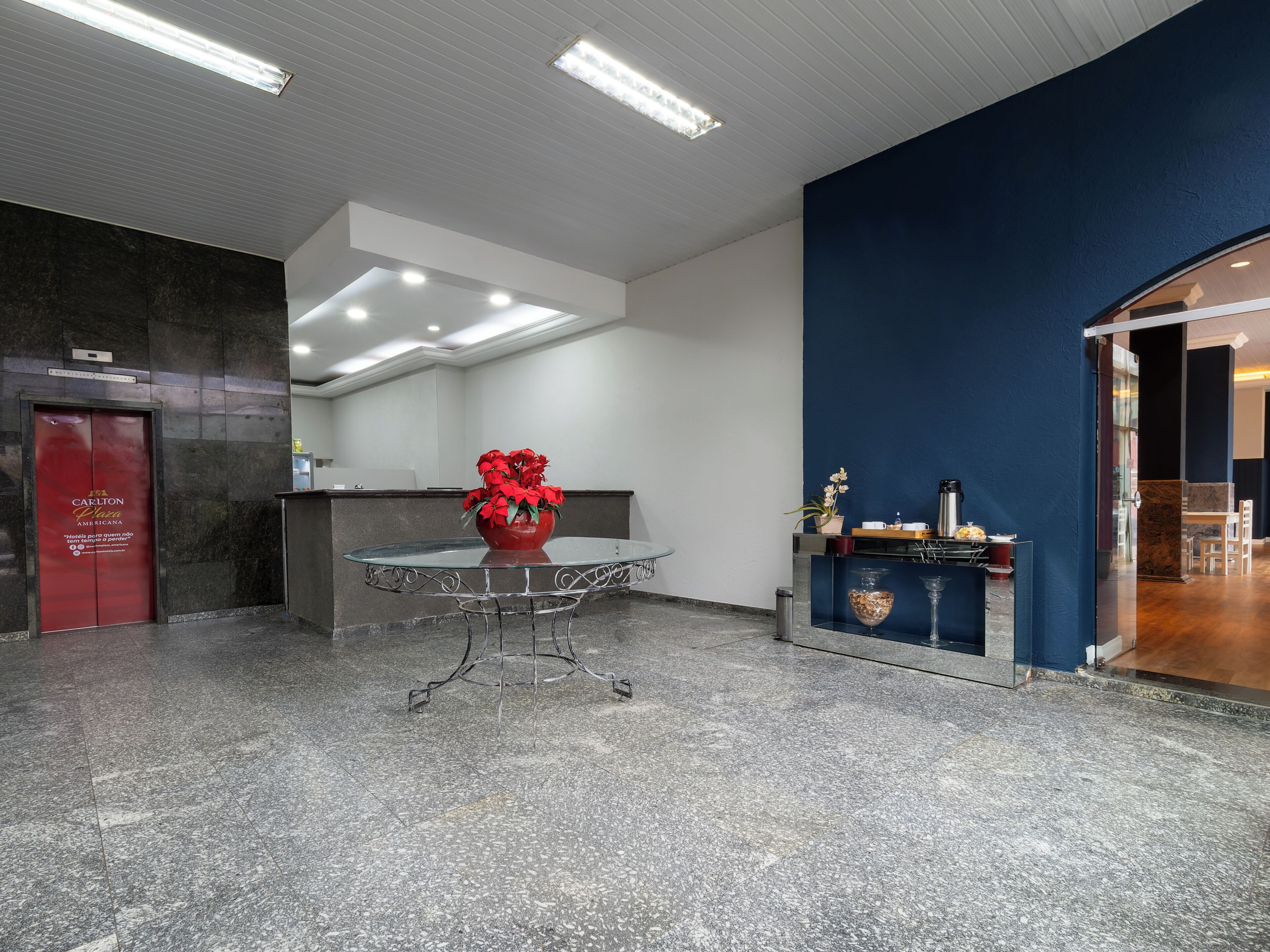 Um hall de hotel com uma mesa de recepção, um vaso de flores vermelho, um espaço para café e uma parede azul escura como detalhe.