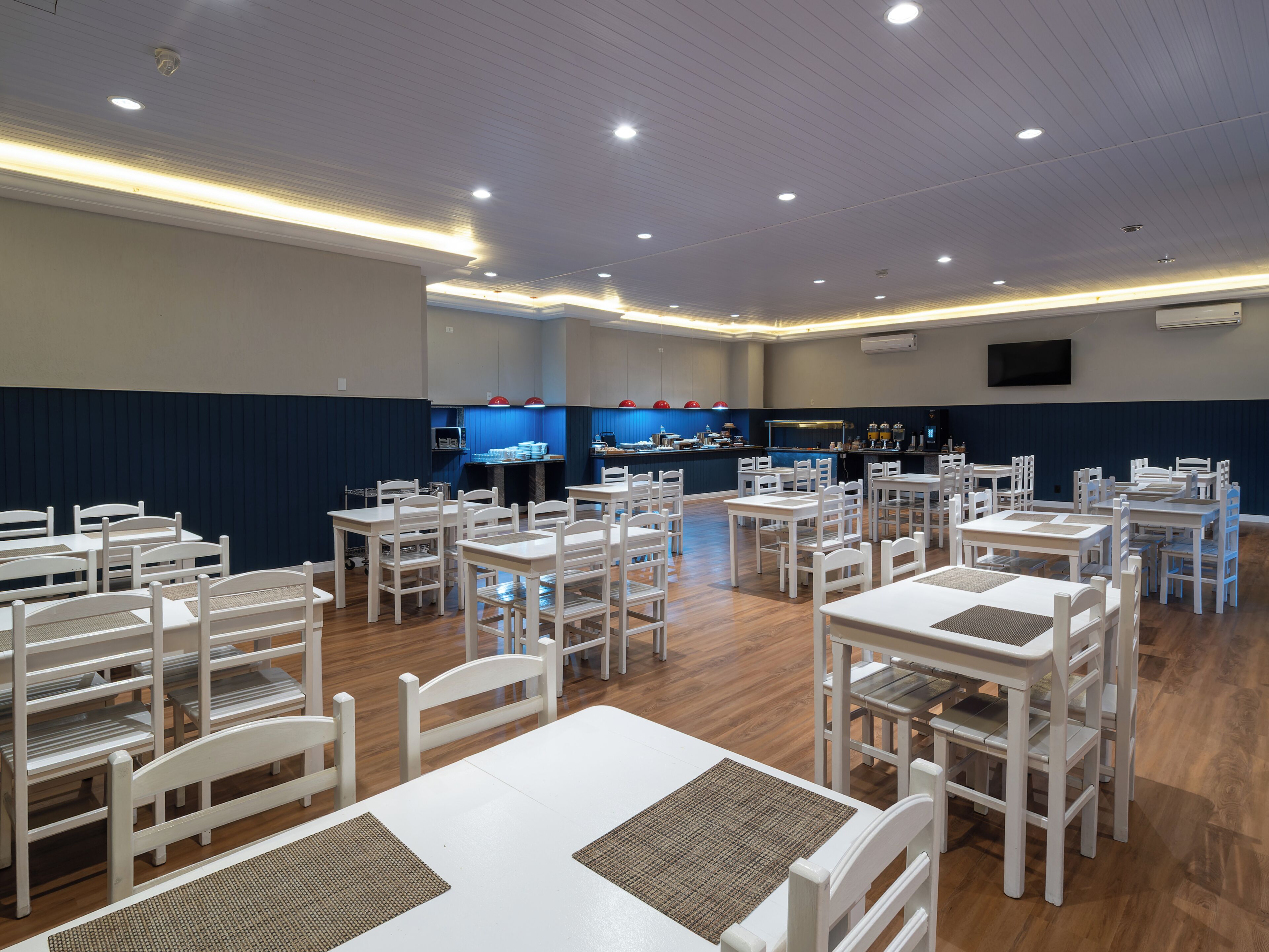 Un restaurante con mesas y sillas blancas, una zona de cocina y un televisor montado en la pared.