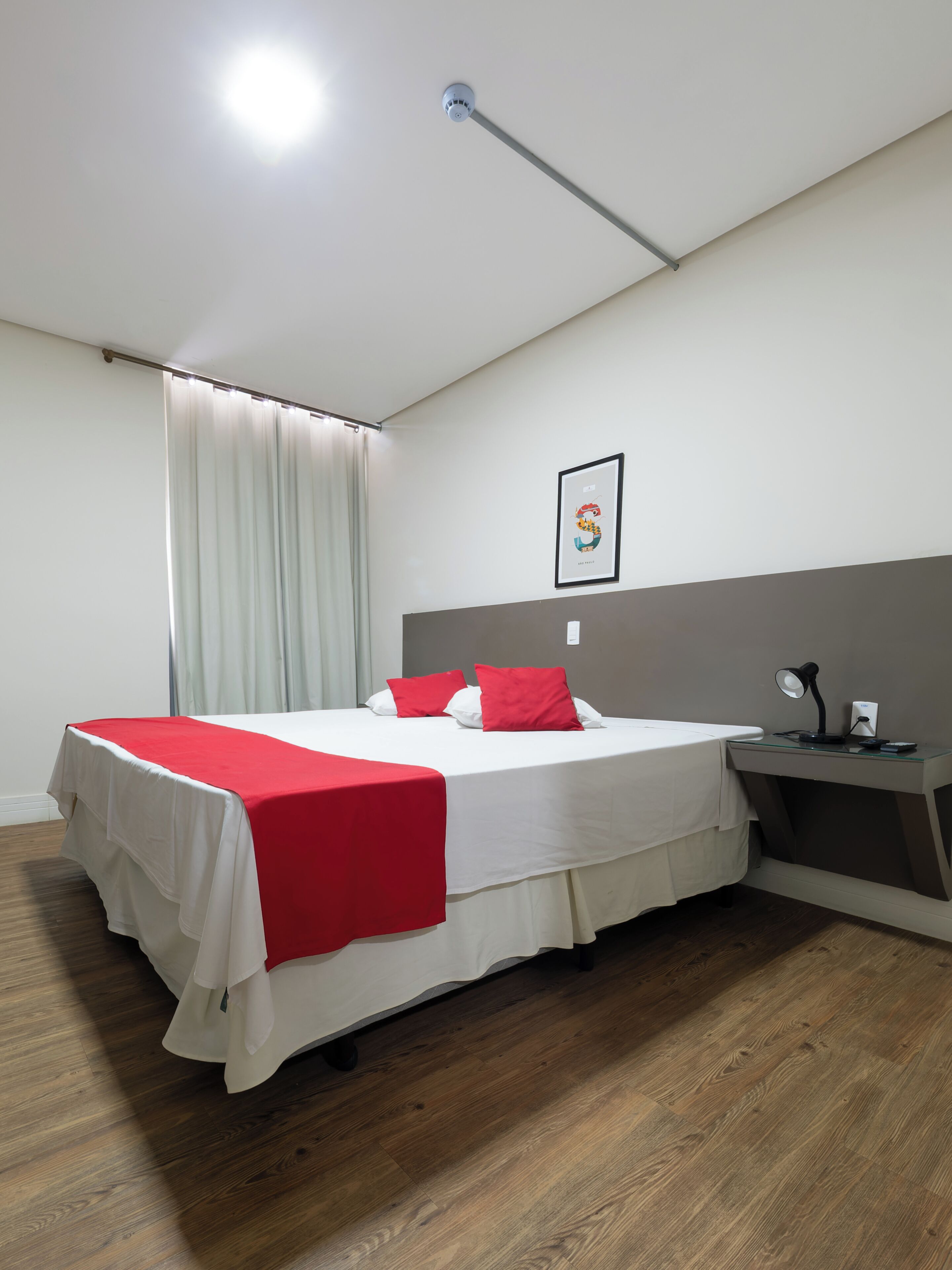 Habitació d'hotel amb llit, escriptori, làmpada, quadre a la paret i finestra amb cortines.