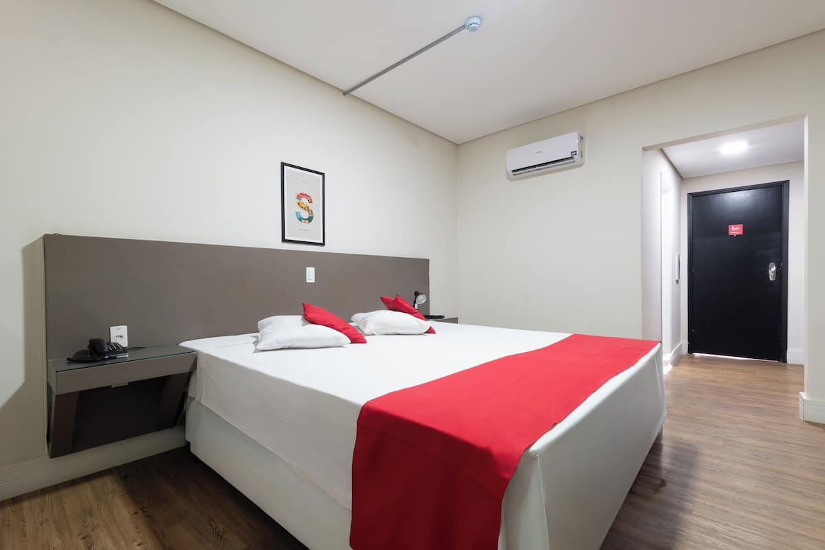 Una camera d'albergo con un letto, un comodino con un telefono, una coperta rossa, un quadro appeso al muro e un condizionatore d'aria.