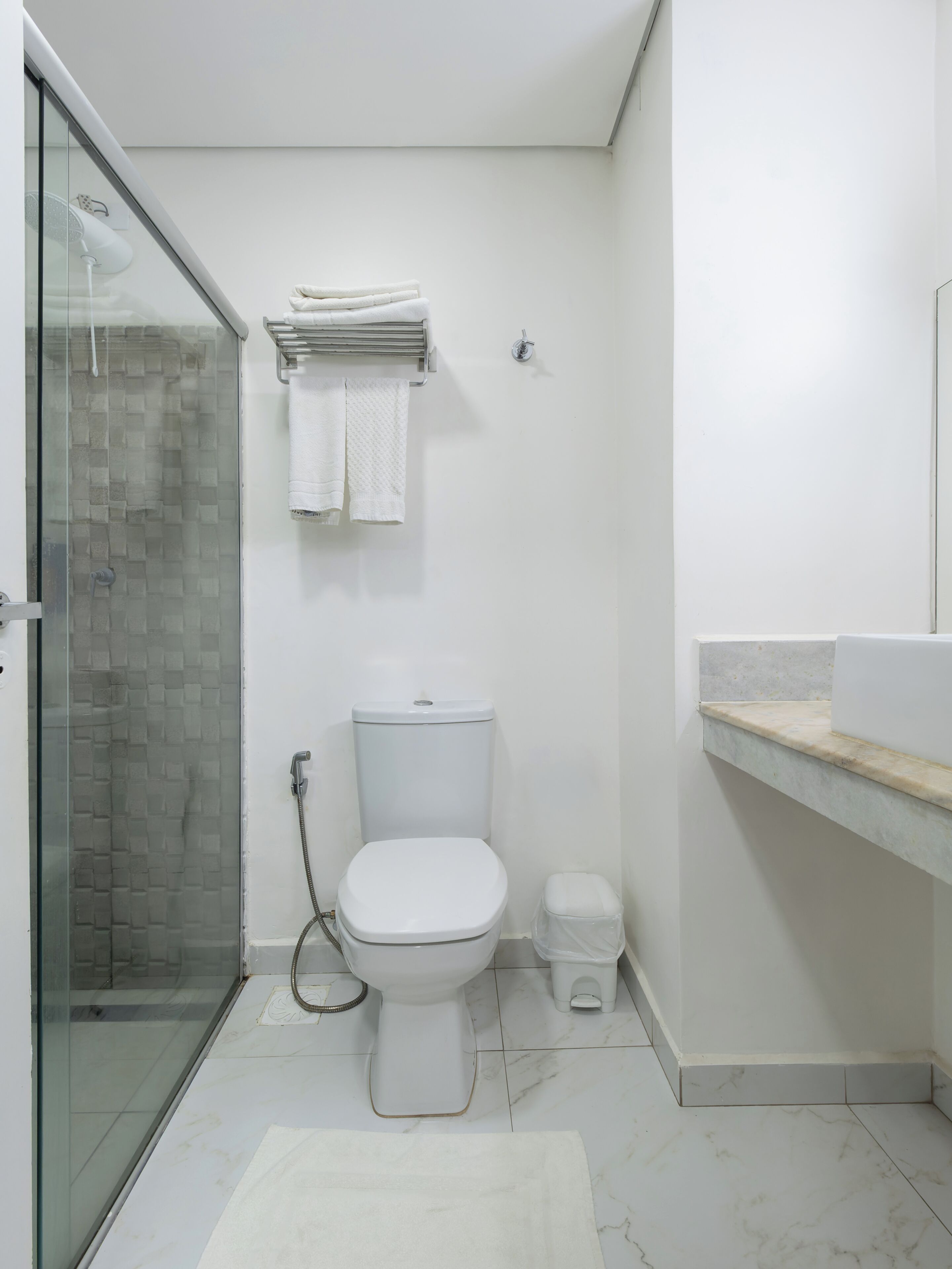 Un bany amb box de dutxa de vidre, un vàter, un lavabo amb taulell de marbre i un terra de rajoles blanques.