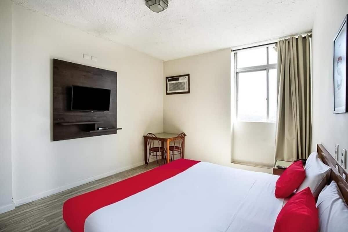 Una camera d'albergo con un letto, una TV a parete, una scrivania con sedie e una finestra con tende.