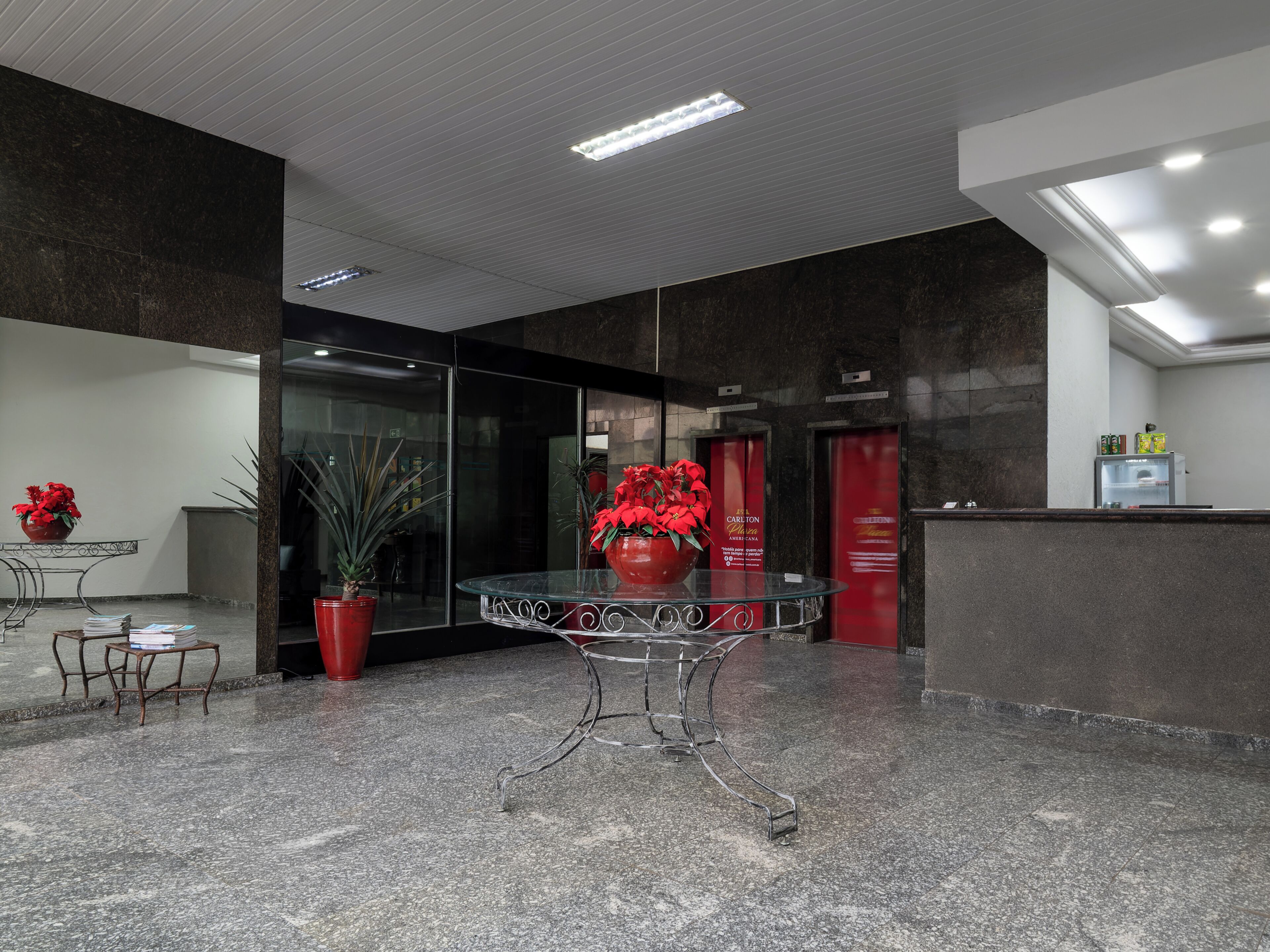 Um hall de entrada com uma mesa de recepção, uma mesa com tampo de vidro e estrutura metálica, uma planta em vaso e um arranjo de flores vermelhas.