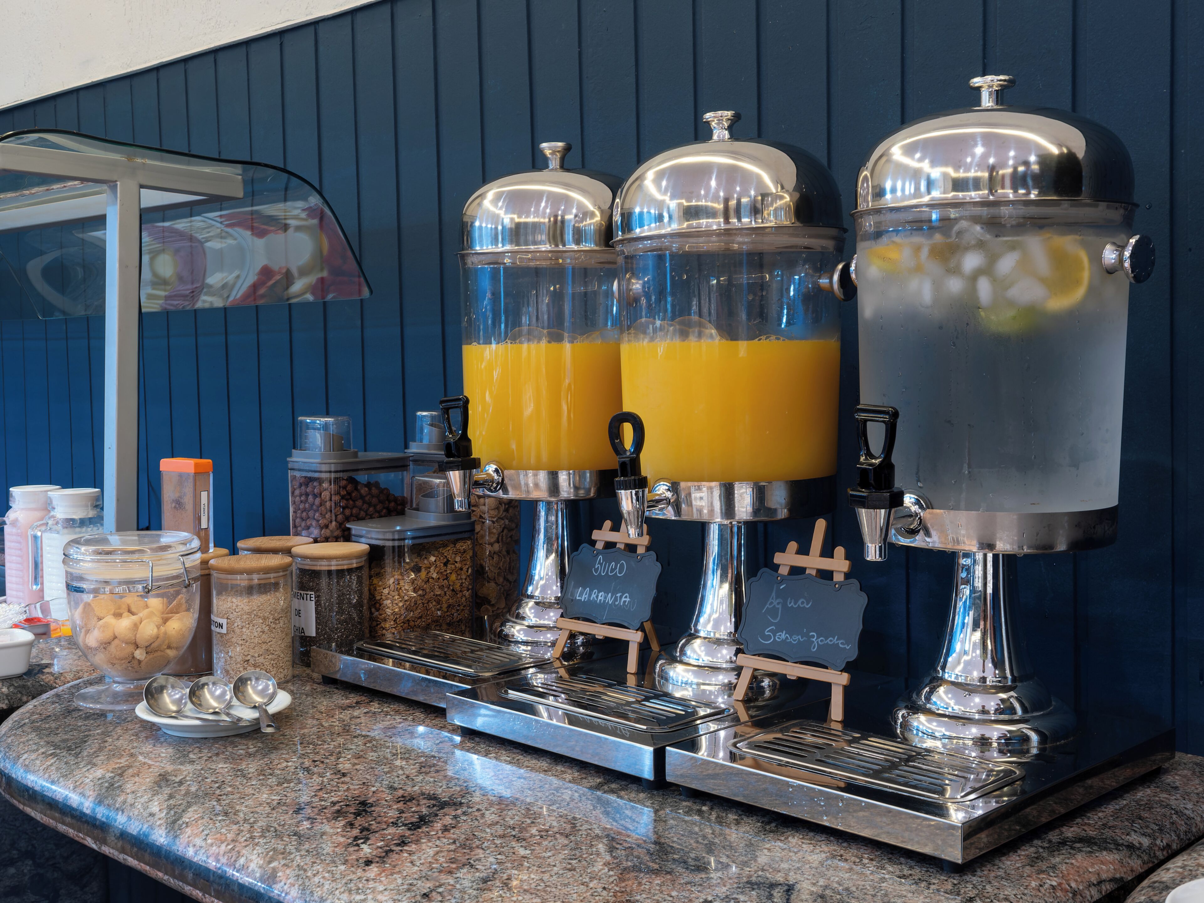 A self-serve beverage station with dispensers for coffee, tea, and water, and a variety of food items on the counter.