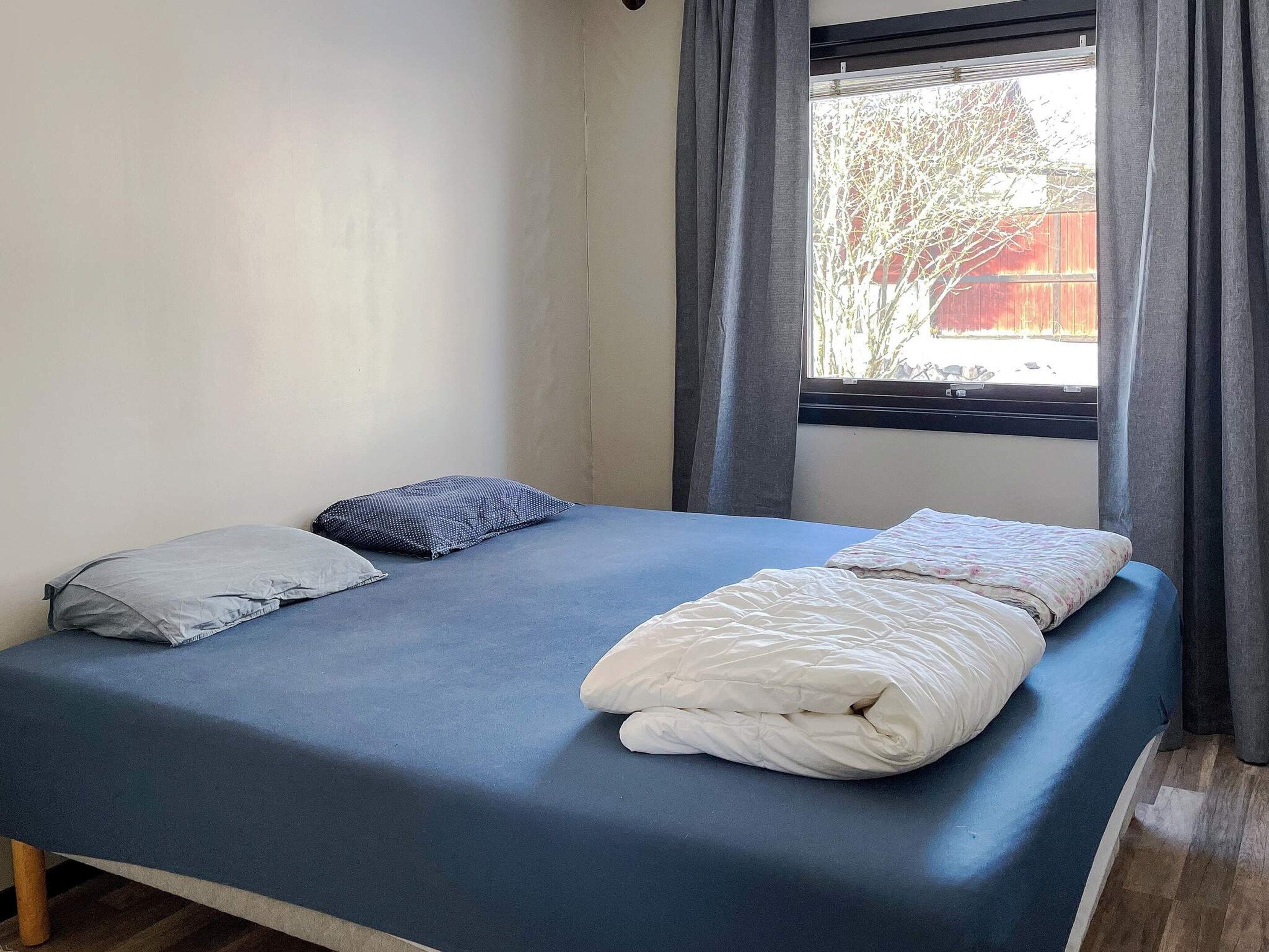 Una cama individual con colchón azul y dos almohadas, una ventana con cortinas y vista a un árbol.