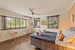 Une chambre à coucher avec un lit, deux chaises, un ventilateur de plafond et de grandes fenêtres équipées de stores.