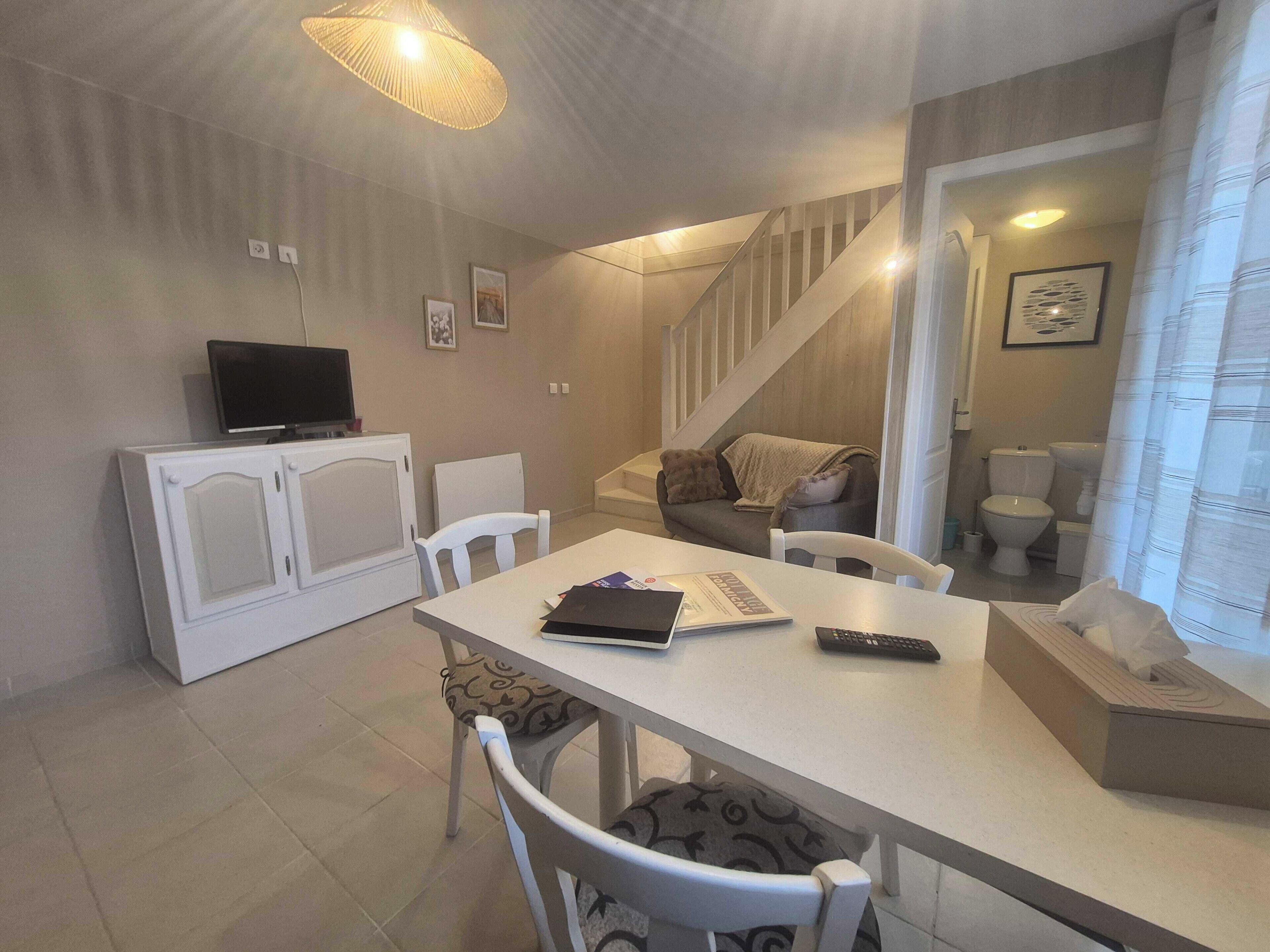 A compact living space with a dining table, chairs, a sofa, a television, and a staircase leading to a bathroom.