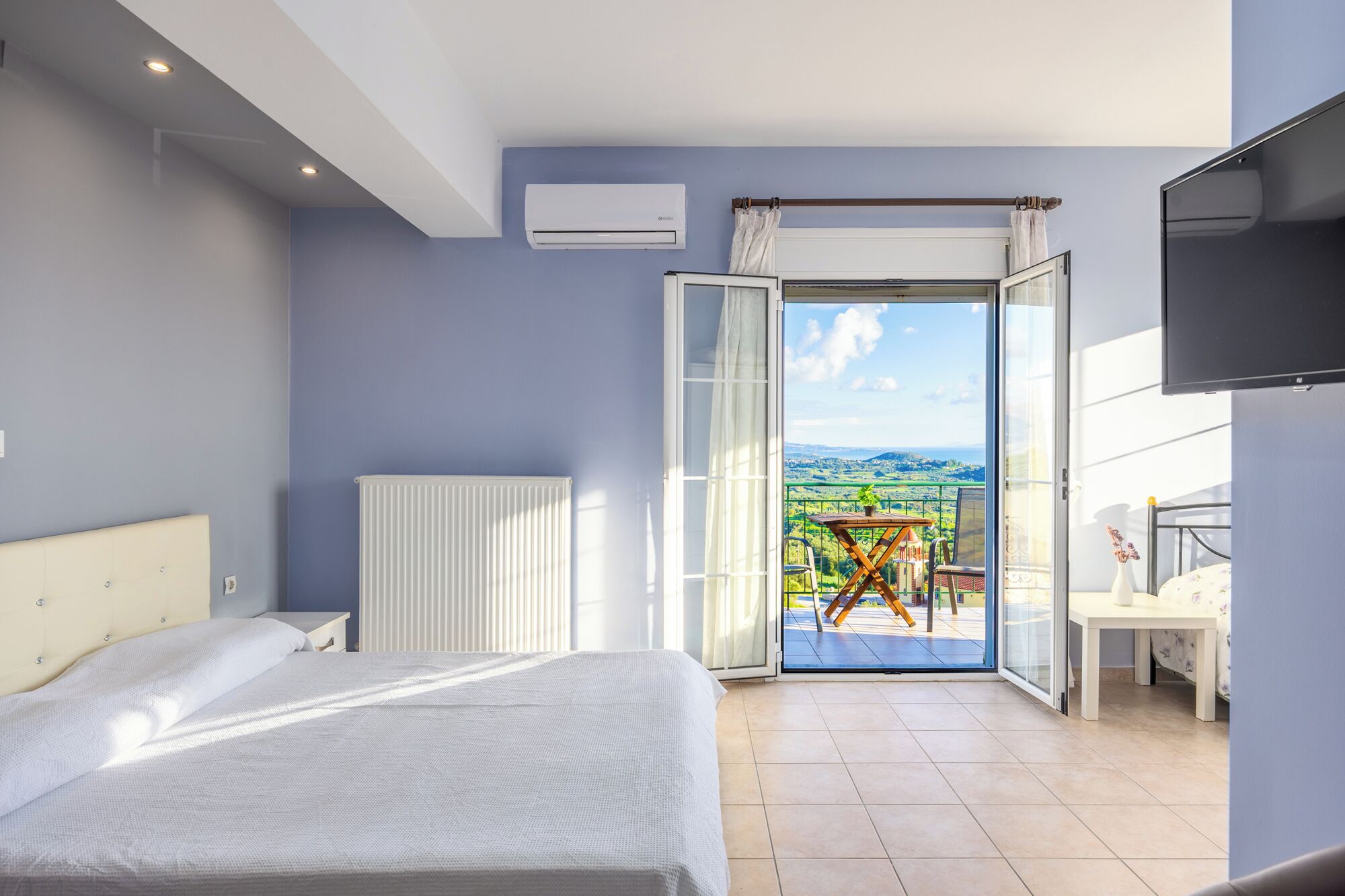 A bedroom with a bed, a radiator, an air conditioning unit, a TV, and a balcony with a table and chairs.