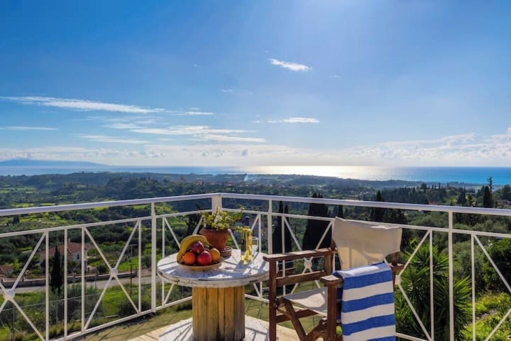 A balcony with a table set with fruits and a potted plant, overlooking a scenic landscape.