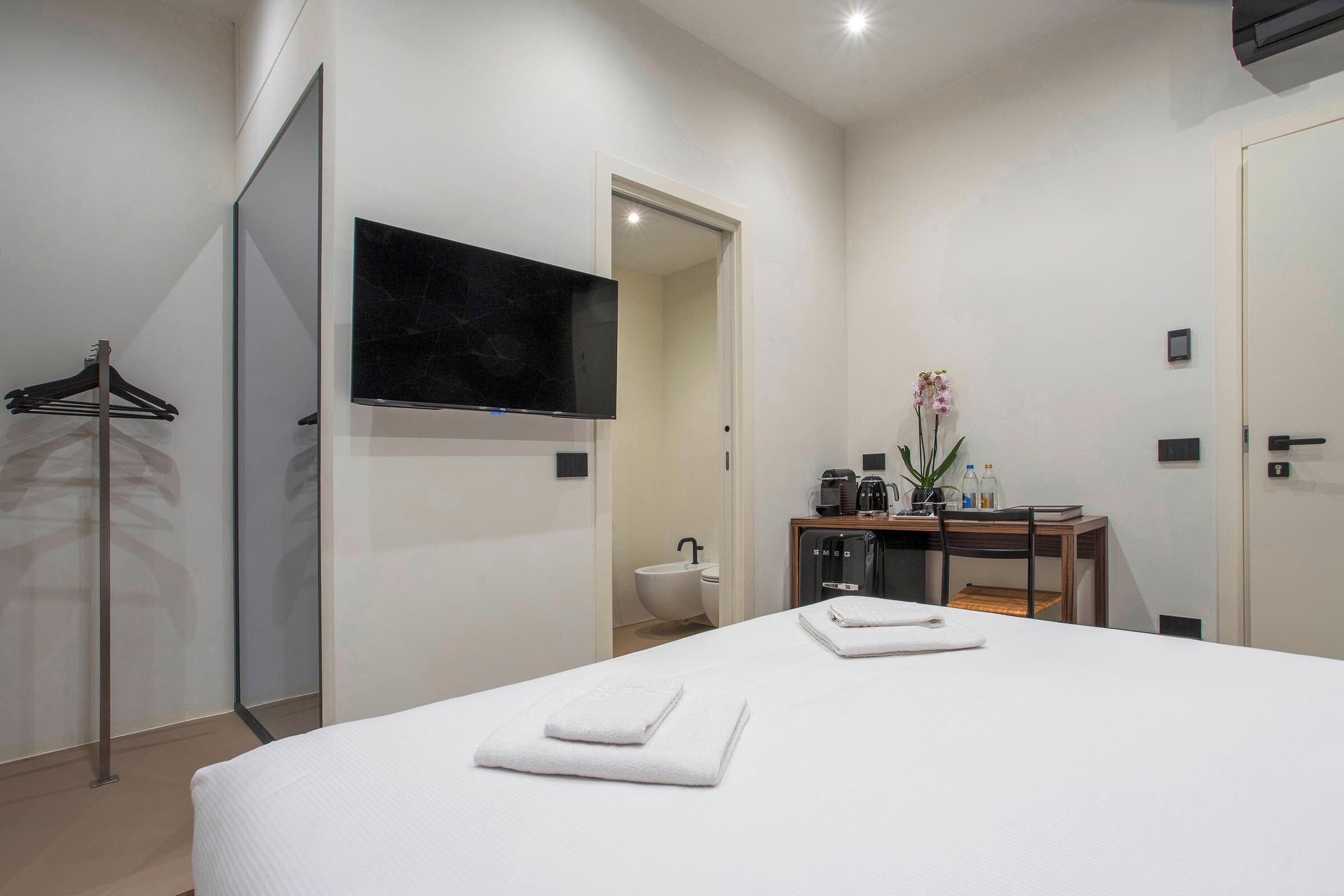 A modern hotel room with a large bed, a flat-screen TV, a desk with a coffee machine, and a bathroom visible through an open door.