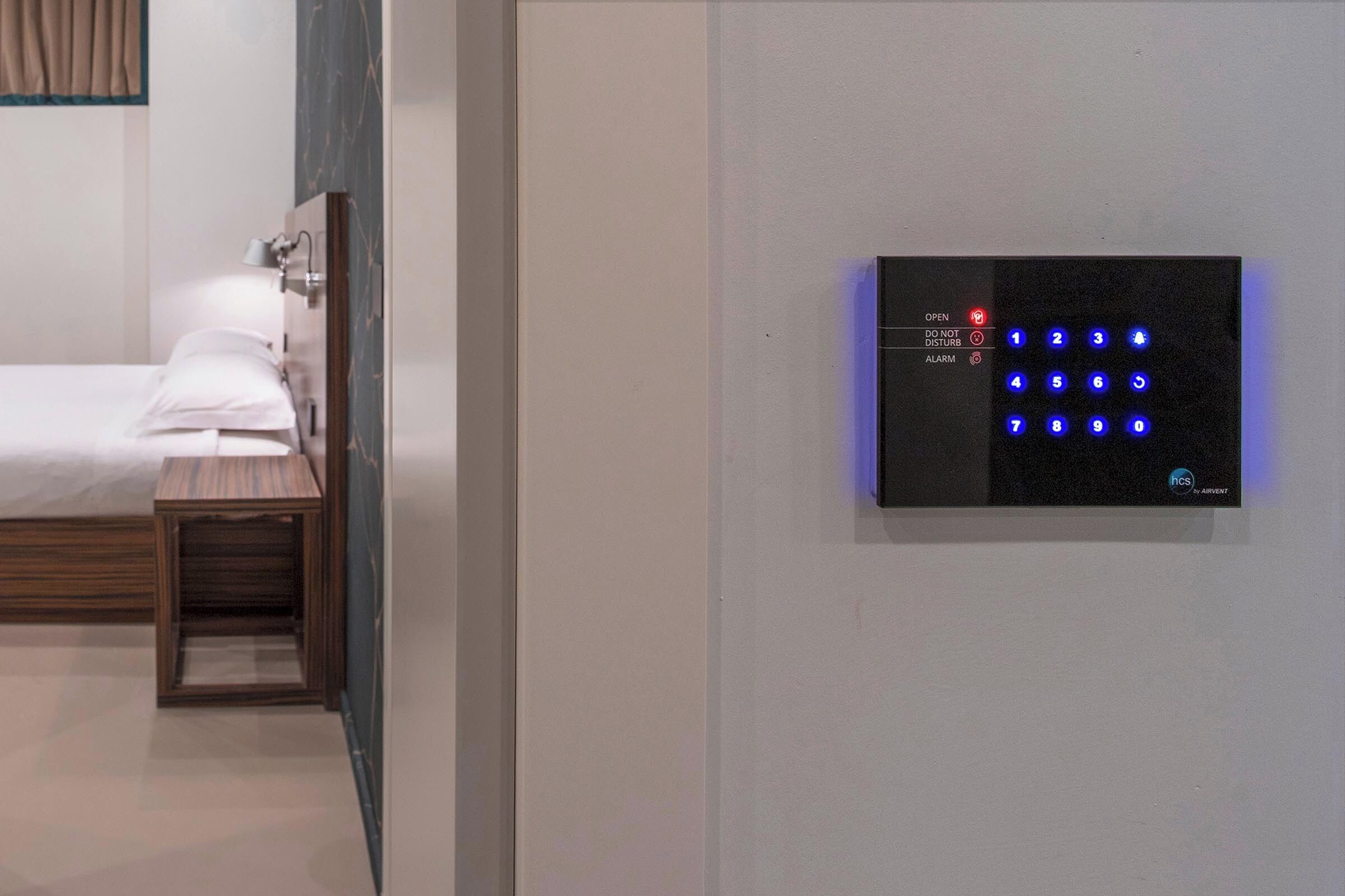 Habitació d'hotel moderna amb llit, taula de nit i teclat electrònic fixat a la paret per accedir-hi.