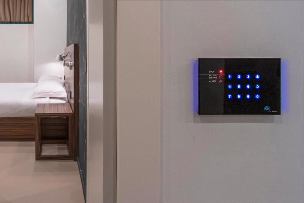 A hotel room with a bed, bedside table, and a wall-mounted electronic keypad for room access.