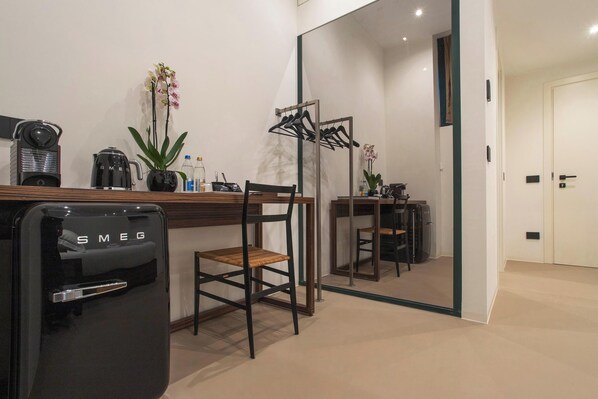 A modern hotel room with a wooden desk, a black Smeg refrigerator, a chair, a small table with a plant, and a mirror reflecting the room. - The Place (Milan)