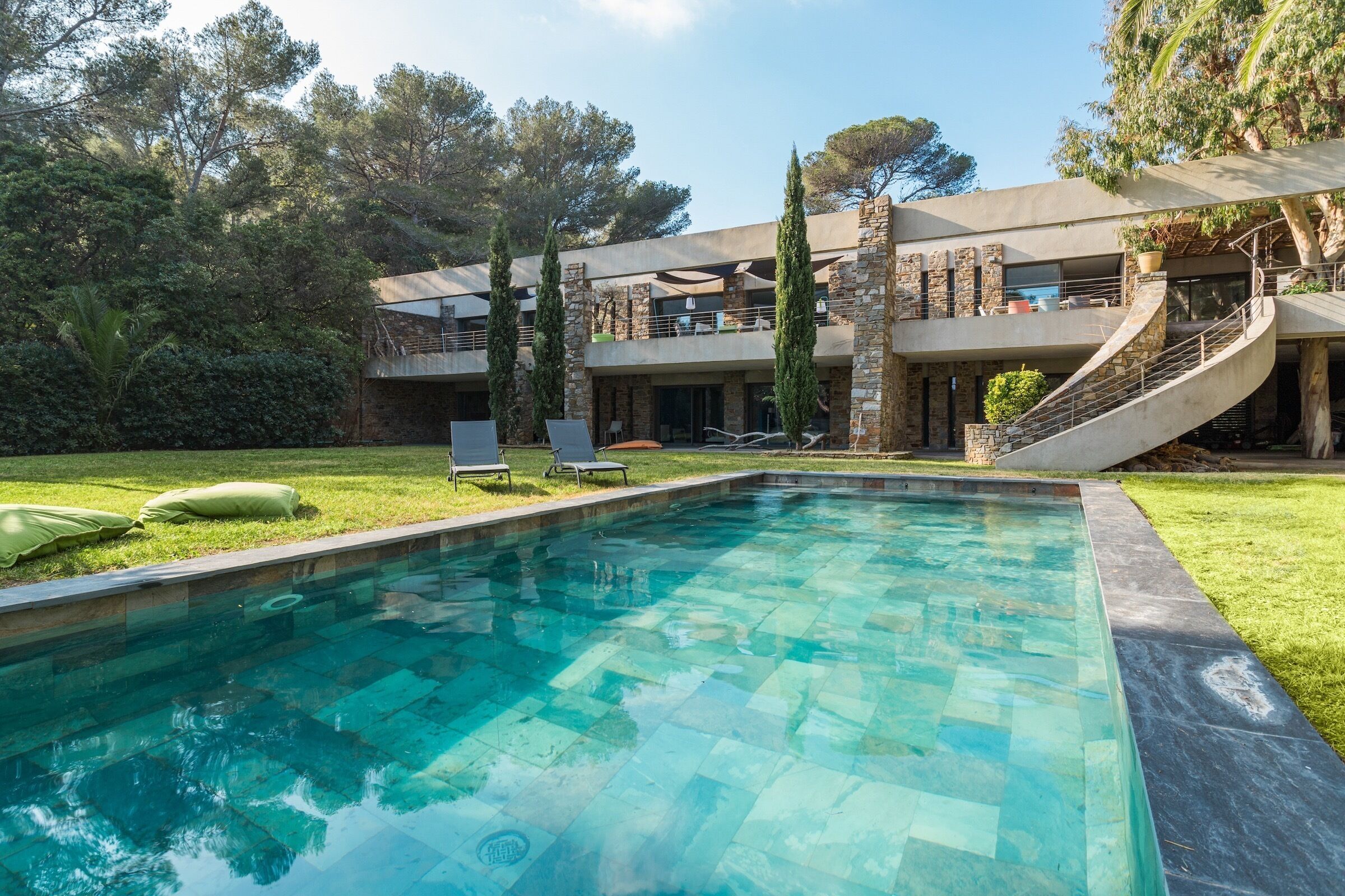 A swimming pool with a stone tile pattern, surrounded by a well-maintained lawn and tall trees.
