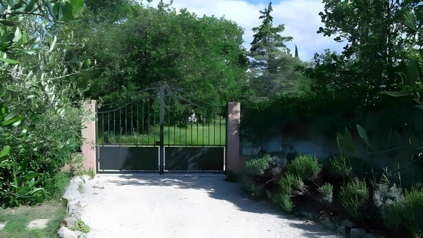Une entrée fermée par un portail en métal, un chemin de gravier et une végétation luxuriante tout autour.