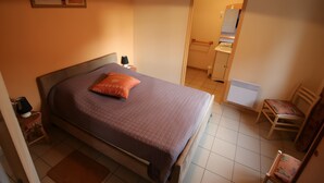 Une chambre à coucher comprenant un lit, une chaise, une table de chevet, une lampe, un tableau accroché au mur et une porte ouverte donnant sur une salle de bain.