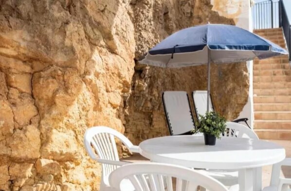 Ein weißer Sitzbereich im Freien mit Tisch und Stühlen, einem blauen Sonnenschirm und einem Blumentopf.