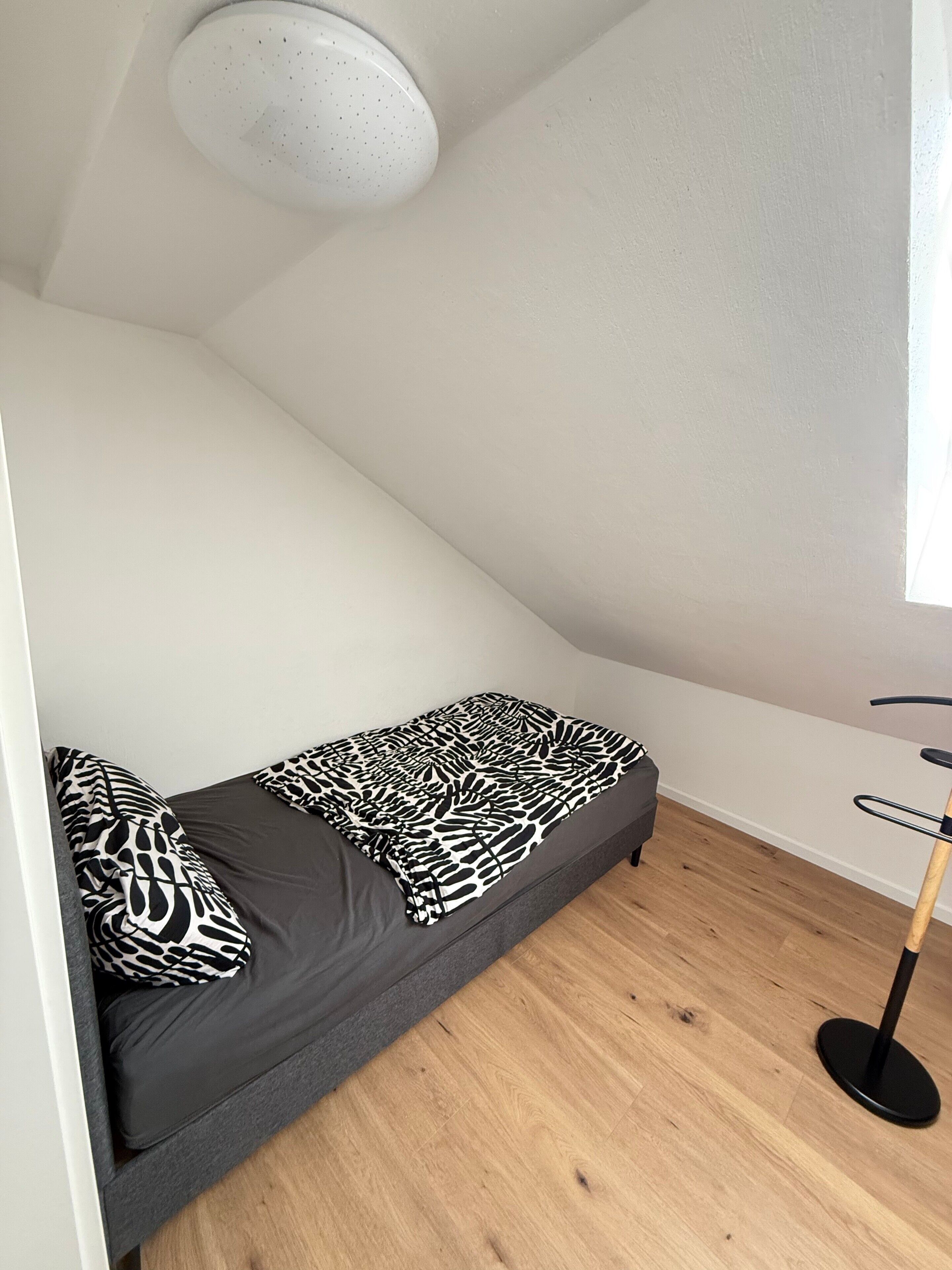 A small, single-bed room with a sloped ceiling, a standing lamp, and a window.