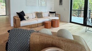 Una sala de estar moderna con sofá, mesa de centro y reposapiés, un ventanal con vistas a zonas verdes, y arte abstracto en la pared.