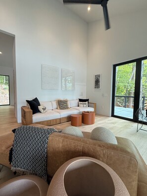 Ein modernes Wohnzimmer mit Sofa, Couchtisch und Hocker, ein großes Fenster mit Blick auf Grünflächen und abstrakte Wandkunst.