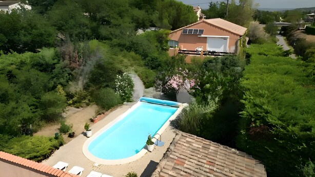 A swimming pool surrounded by greenery, a house with a garage, and a paved area with lounge chairs. - Gite la Roseraie with Pool (Ruoms)