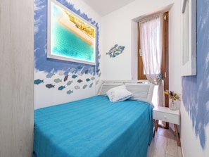 Un dormitorio con cama, una ventana que muestra una escena de playa, una pared decorada con motivos de peces y una mesita con un jarrón.