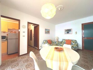 A dining area with a round table, chairs, a patterned floor, a kitchen with a refrigerator, and a hallway leading to another room. - Sunny Beachside Apartment in Grado (Grado)