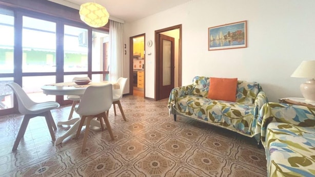 Ein Wohnzimmer mit gemustertem Boden, einer Couch mit floralem Bezug, einem Essbereich mit Tisch und Stühlen sowie einer durchgehenden Tür erkennbaren Küche.