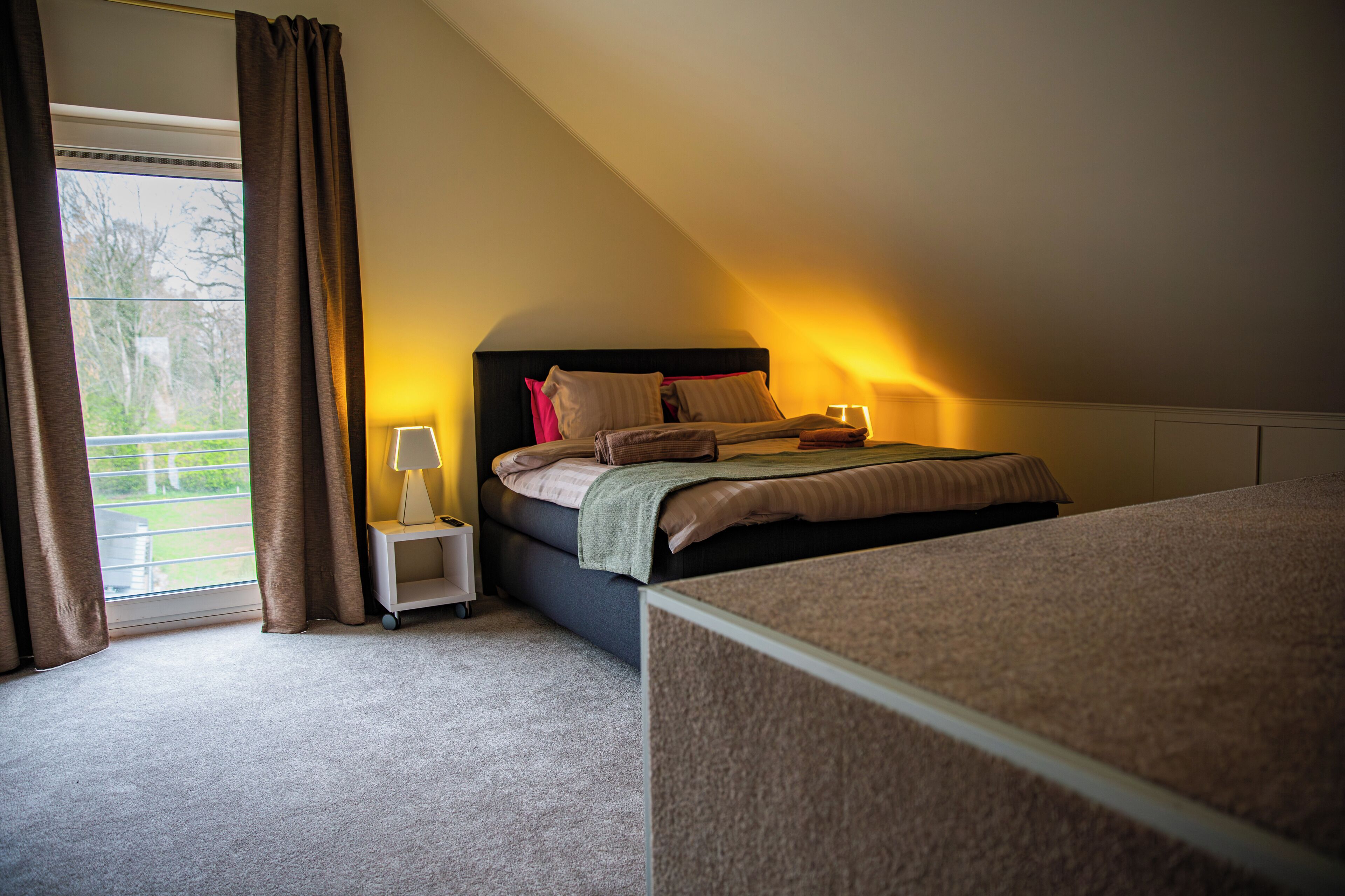 A bedroom with a bed, a nightstand, a lamp, and a view of the outdoors.