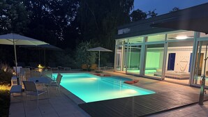Un área de piscina moderna al aire libre con sillones de descanso y sombrillas, un spa con paredes de vidrio y una terraza de madera.