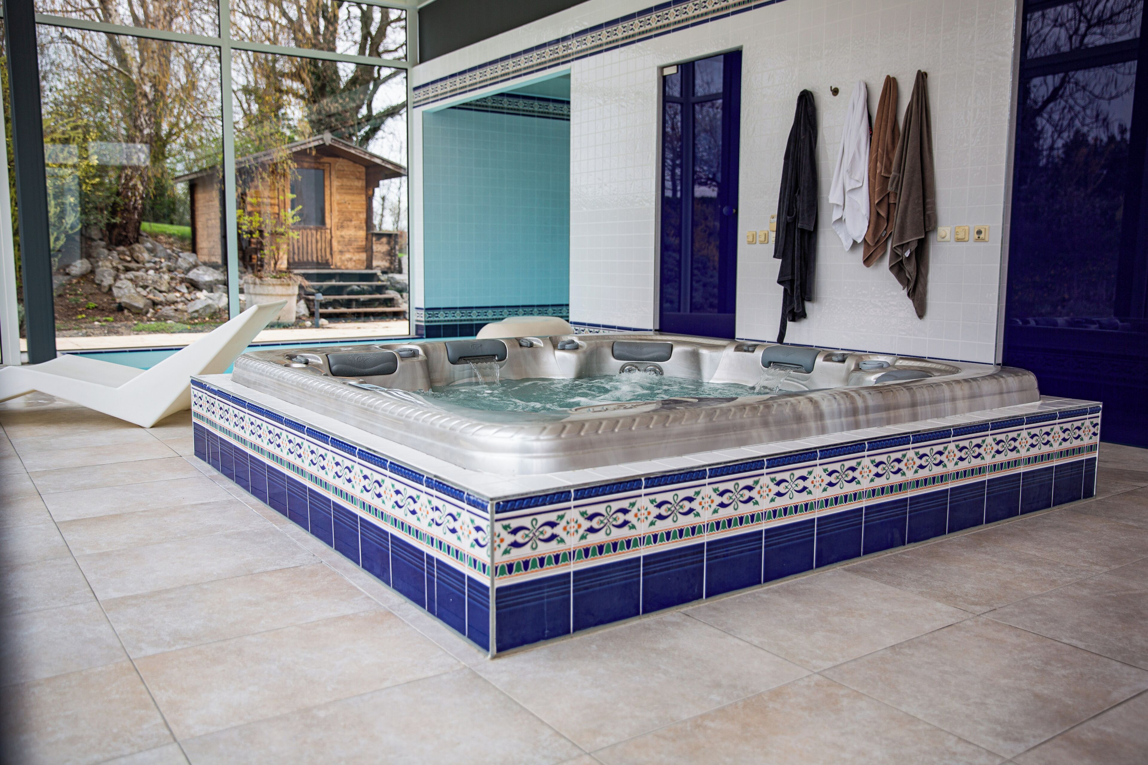 Ein Whirlpool mit gefliestem Beckenrand, einer Holzterrasse und Blick auf ein Haus durch eine Glastür.