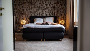 Una cama bien hecha con almohadas y una toalla enrollada, un papel tapiz estampado y una lámpara de noche.