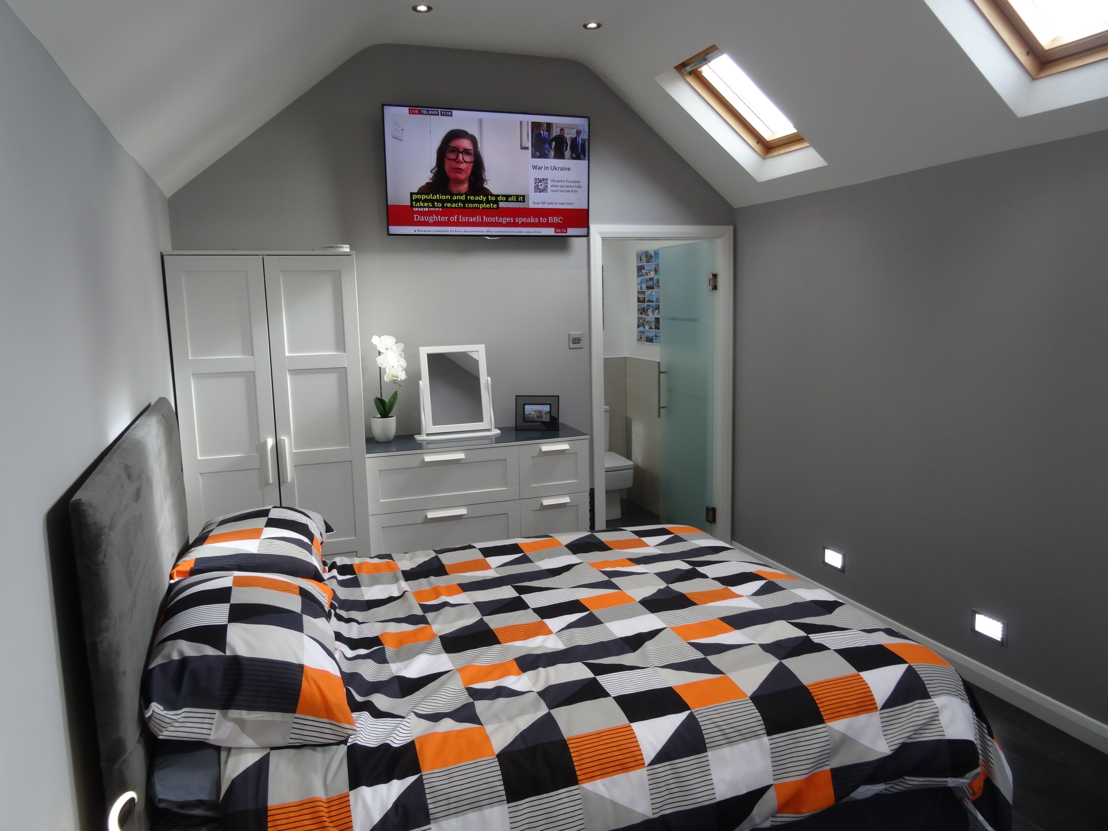 Un dormitorio con una cama, un aparador, un espejo y un televisor que muestra un noticiero.