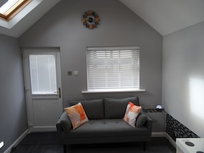 A room with a grey sofa, two windows with blinds, a door, a wall clock, and a skylight.